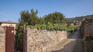 Un estrecho sendero de piedra flanqueado por un muro de piedra y una puerta de madera, que conduce a una zona residencial con casas y vegetación.