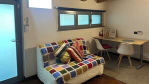 Una habitación acogedora con techo de madera, una ventana con persianas, un sofá colorido de ganchillo, una silla blanca y un escritorio con un ordenador.