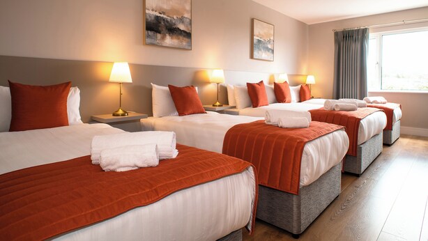 Una camera d'albergo con quattro letti, ciascuno con una coperta rossa e cuscini bianchi, e un'ampia finestra con tende.