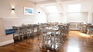 Zona pranzo con diversi tavoli e sedie, soffitto bianco con lucernari e un quadro appeso al muro.