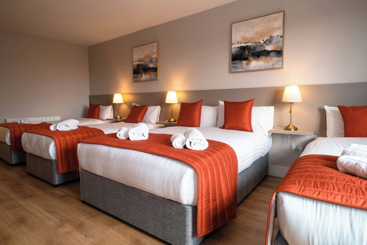 A hotel room with four beds, each with a red blanket and white pillows, and a bedside table with a lamp.