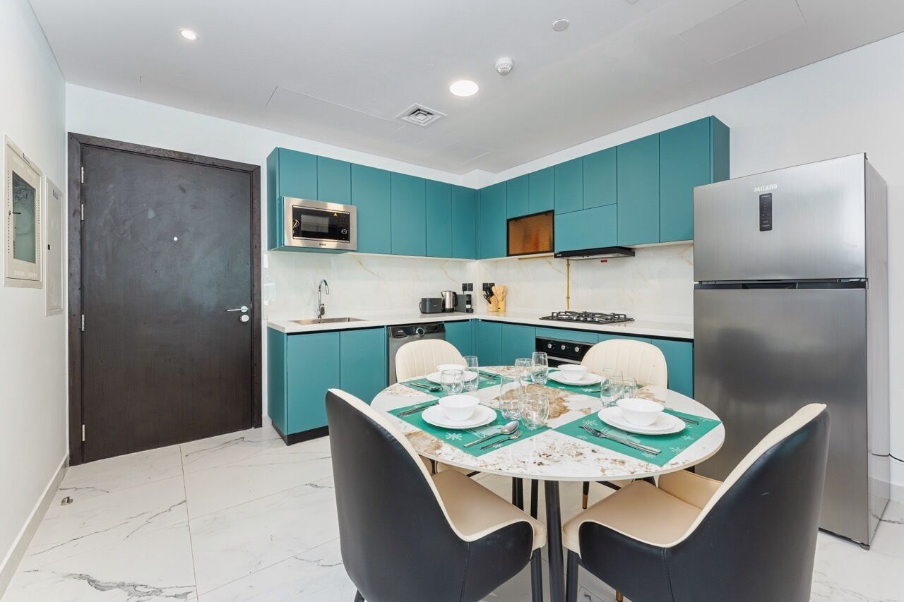 A modern kitchen with teal cabinets, a stainless steel refrigerator, and a dining table set for four.