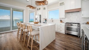 A modern kitchen with a large island, wooden bar stools, a stainless steel refrigerator, and a view of the ocean.