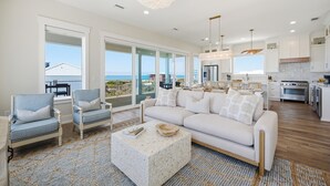 A spacious living area with a large sofa, armchairs, and a coffee table. There is a kitchen with modern appliances and a view of the ocean.