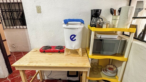 A small kitchen area with a wooden table, a red gas stove, a white container with a blue lid, a yellow shelf with kitchen utensils, and a red patterned floor. - Apto Maison | 5-minute walk from Central Park (Antigua Guatemala)