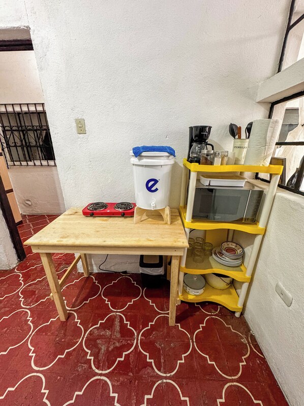 A small kitchen area with a wooden table, a red gas stove, a white container with a blue lid, a yellow shelf with kitchen utensils, and a red patterned floor.