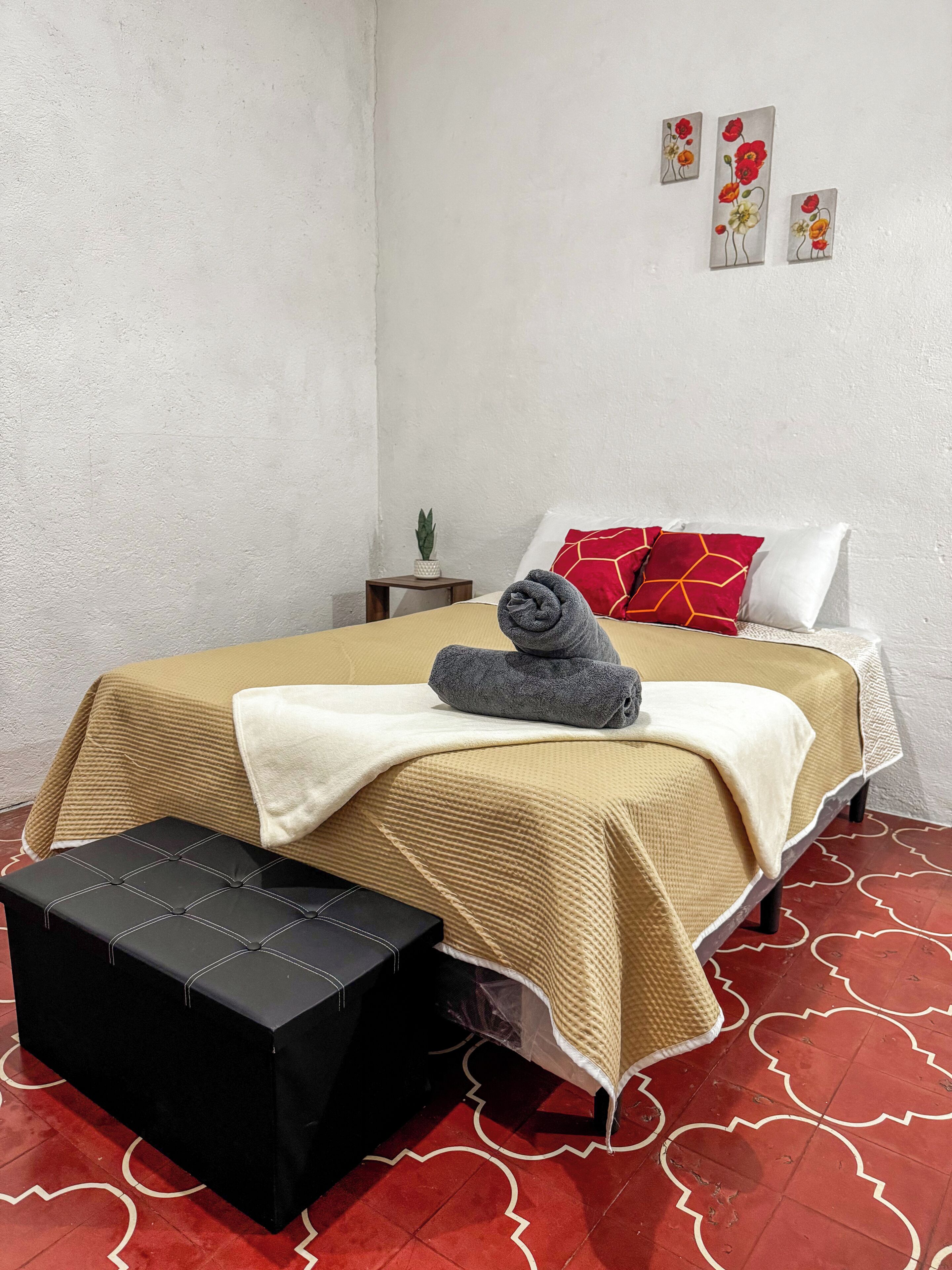 A neatly made bed with a beige cover, a rolled towel, and a red pillow. There is a small bedside table with a plant, and a black bench with a white cushion. The floor has a red pattern.