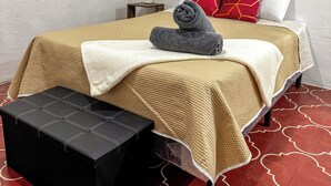 A neatly made bed with a beige cover, a rolled towel, and a red pillow. There is a small bedside table with a plant, and a black bench with a white cushion. The floor has a red pattern. - Apto Maison | 5-minute walk from Central Park (Antigua Guatemala)
