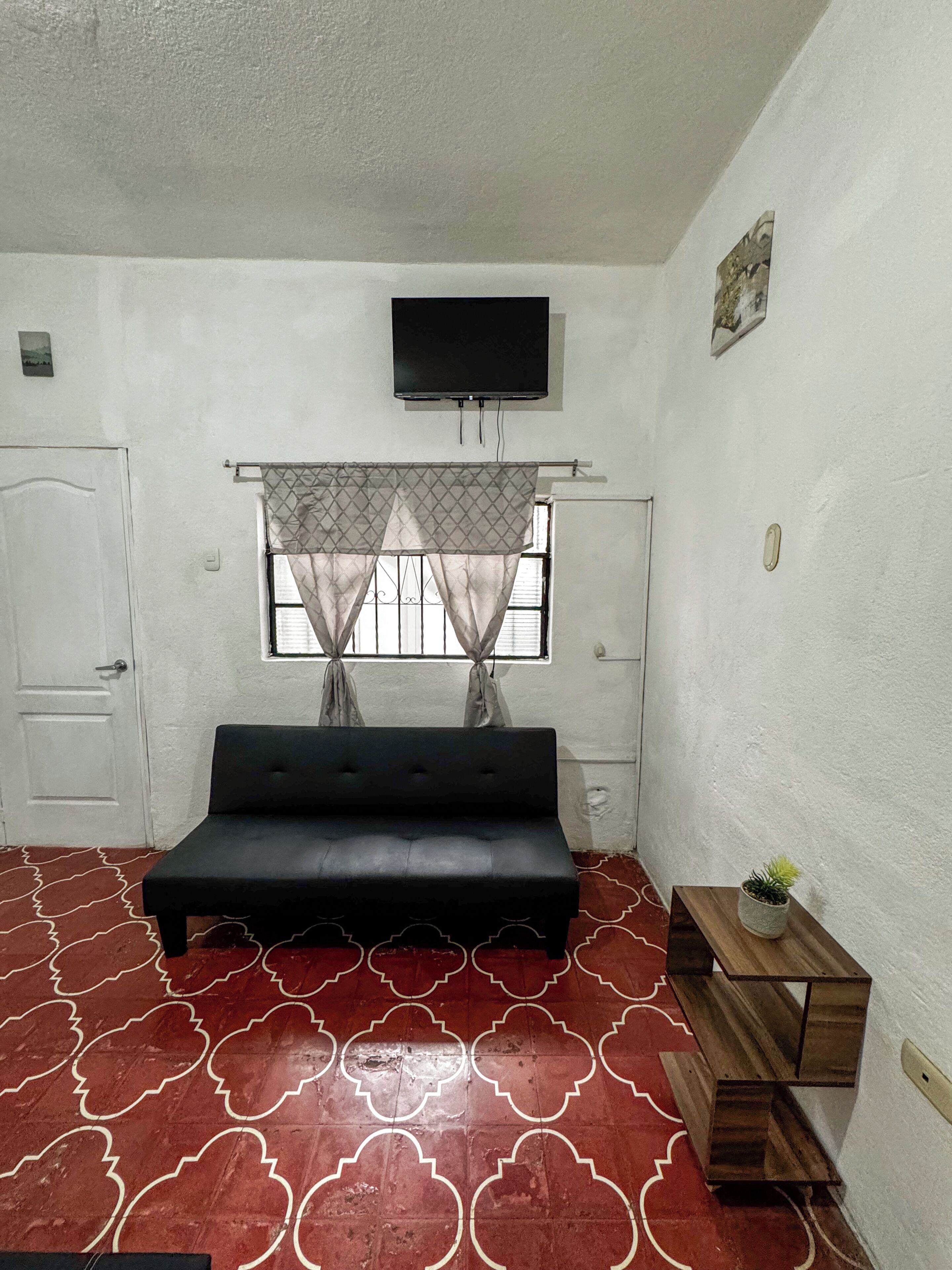 Una habitación con una cama, televisión montada en la pared, ventana con cortinas y una pequeña mesa con una planta.