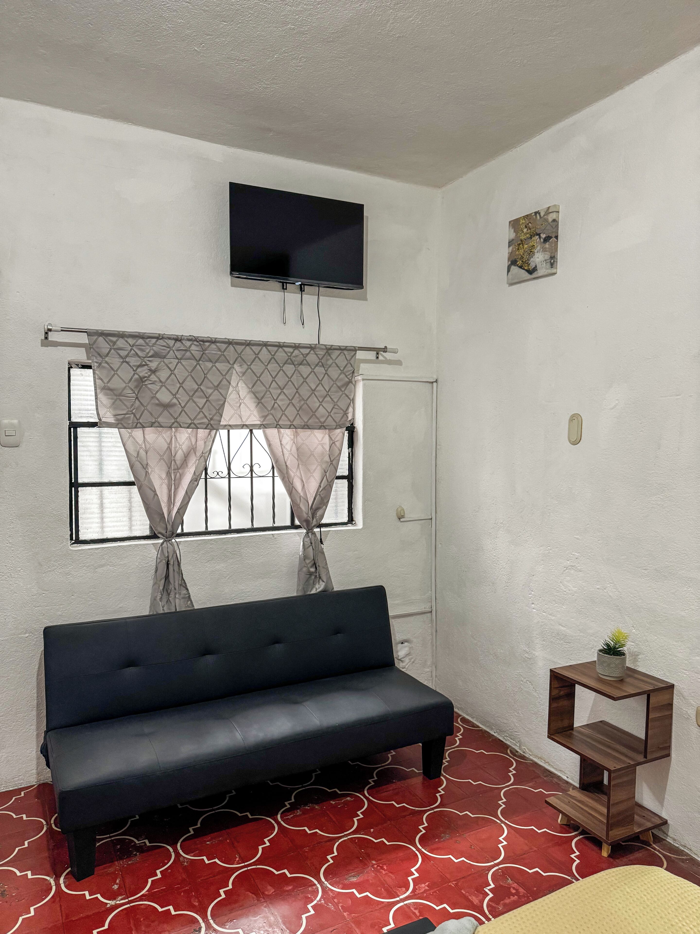 A room with a sofa, a television mounted on the wall, a window with curtains, and a small wooden shelf with a plant.