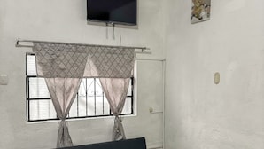 A room with a sofa, a television mounted on the wall, a window with curtains, and a small wooden shelf with a plant. - Apto Maison | 5-minute walk from Central Park (Antigua Guatemala)