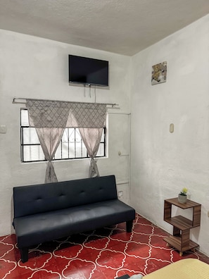 A room with a sofa, a television mounted on the wall, a window with curtains, and a small wooden shelf with a plant.