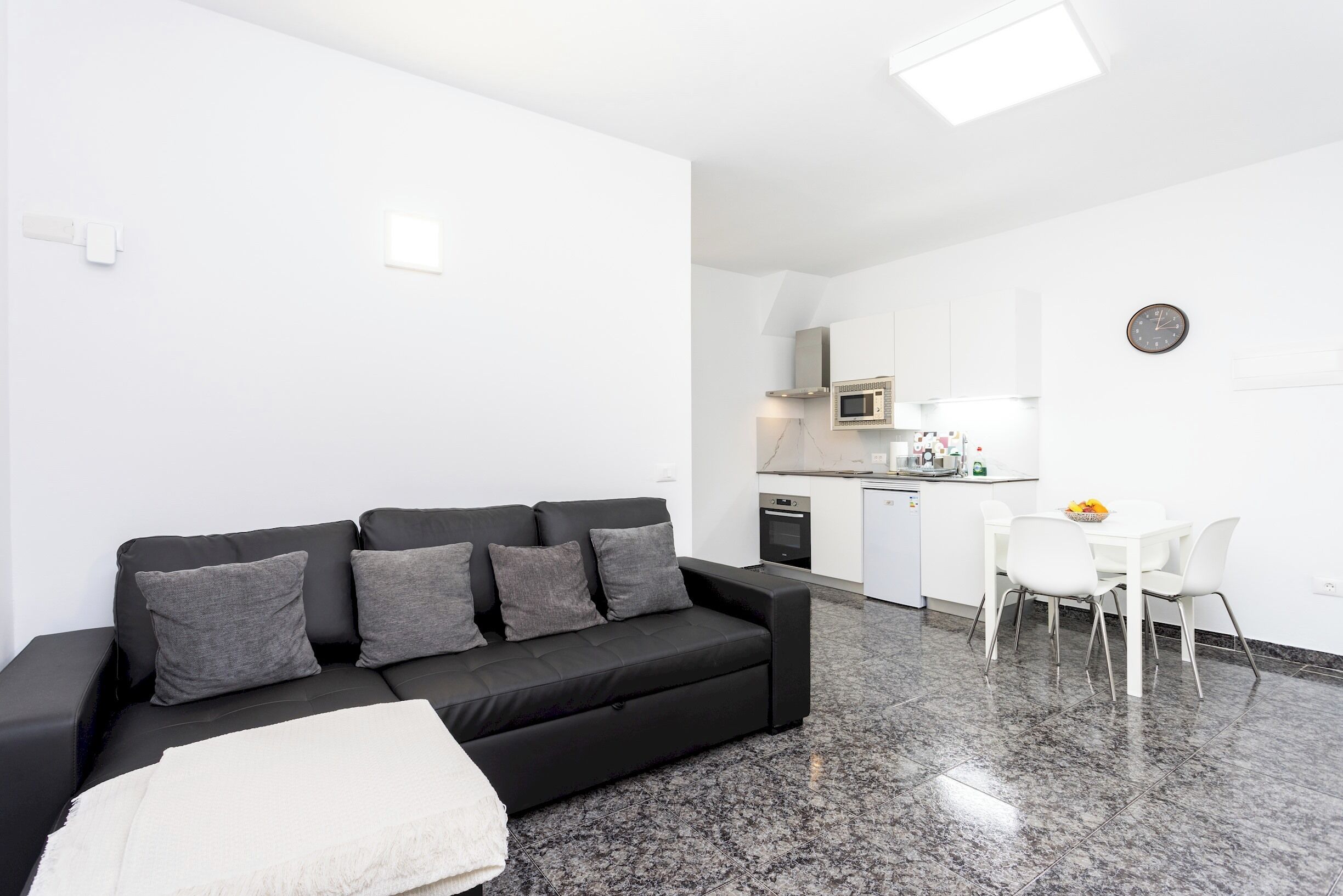 Uma sala de estar moderna com um sofá preto, uma mesa de jantar branca com cadeiras, uma área de cozinha com armários brancos e um relógio na parede.