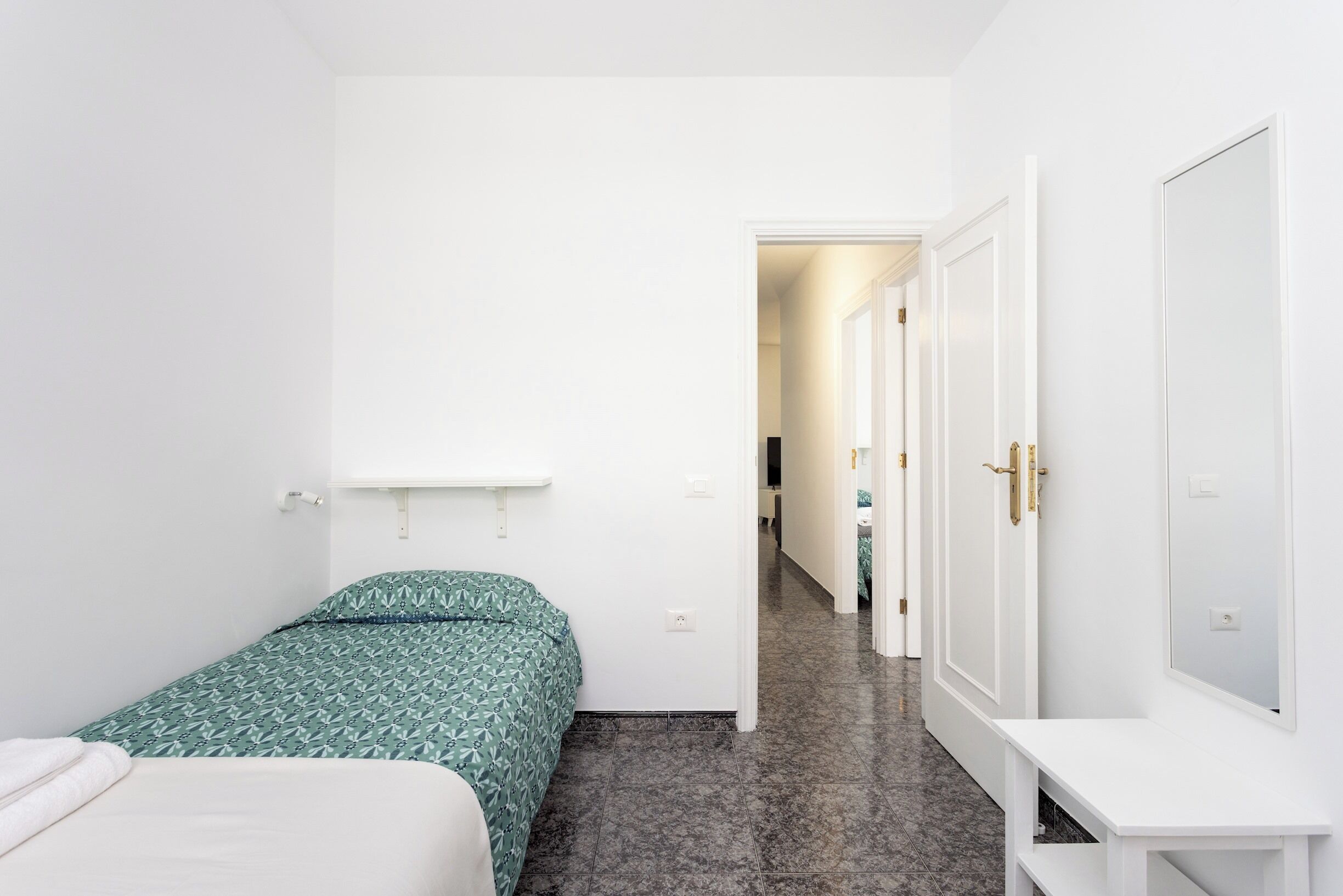 Un couloir étroit mène à une chambre à coucher qui contient un lit, un banc blanc et une armoire avec un miroir.