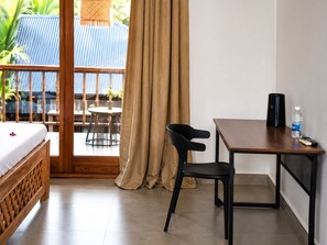 A hotel room with a bed, a desk, a chair, a small table with a plant, and a view of a blue-roofed building and palm trees. - The Lagoons Turtle Resort (Nungwi)