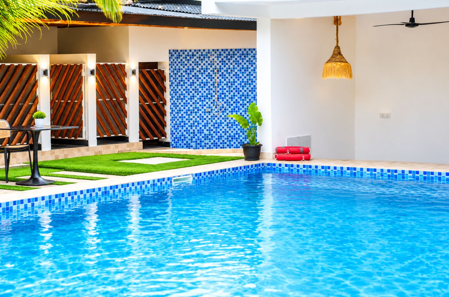 Une piscine dont le bord est recouvert de carreaux bleus, un petit jardin avec de l’herbe et une plante en pot, et une clôture en bois avec des lattes disposées en diagonale.