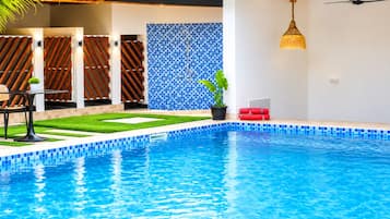 Une piscine dont le bord est recouvert de carreaux bleus, un petit jardin avec de l’herbe et une plante en pot, et une clôture en bois avec des lattes disposées en diagonale.
