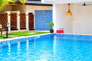 Una piscina con borde de azulejos azules, un pequeño jardín con césped y una planta en maceta, y una valla de madera con listones diagonales.