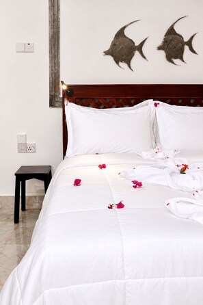 A neatly made bed with white linens and scattered rose petals, a wooden headboard, and two wall-mounted fish decorations. - The Lagoons Turtle Resort (Nungwi)