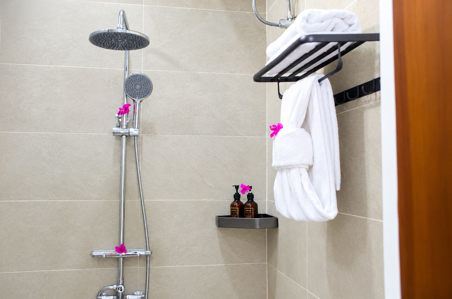 Een douche met een handdouche, een in de wand gemonteerde regendouche en een handdoekrek met een handdoek en een roze bloem.