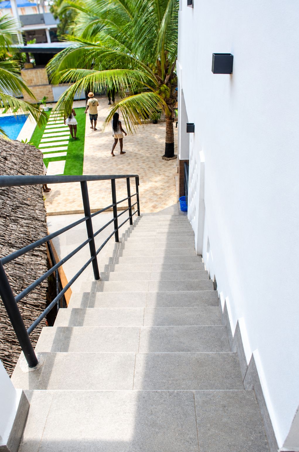 Un escalier menant à un bâtiment blanc doté d’une rampe métallique, un palmier et un ciel dégagé.