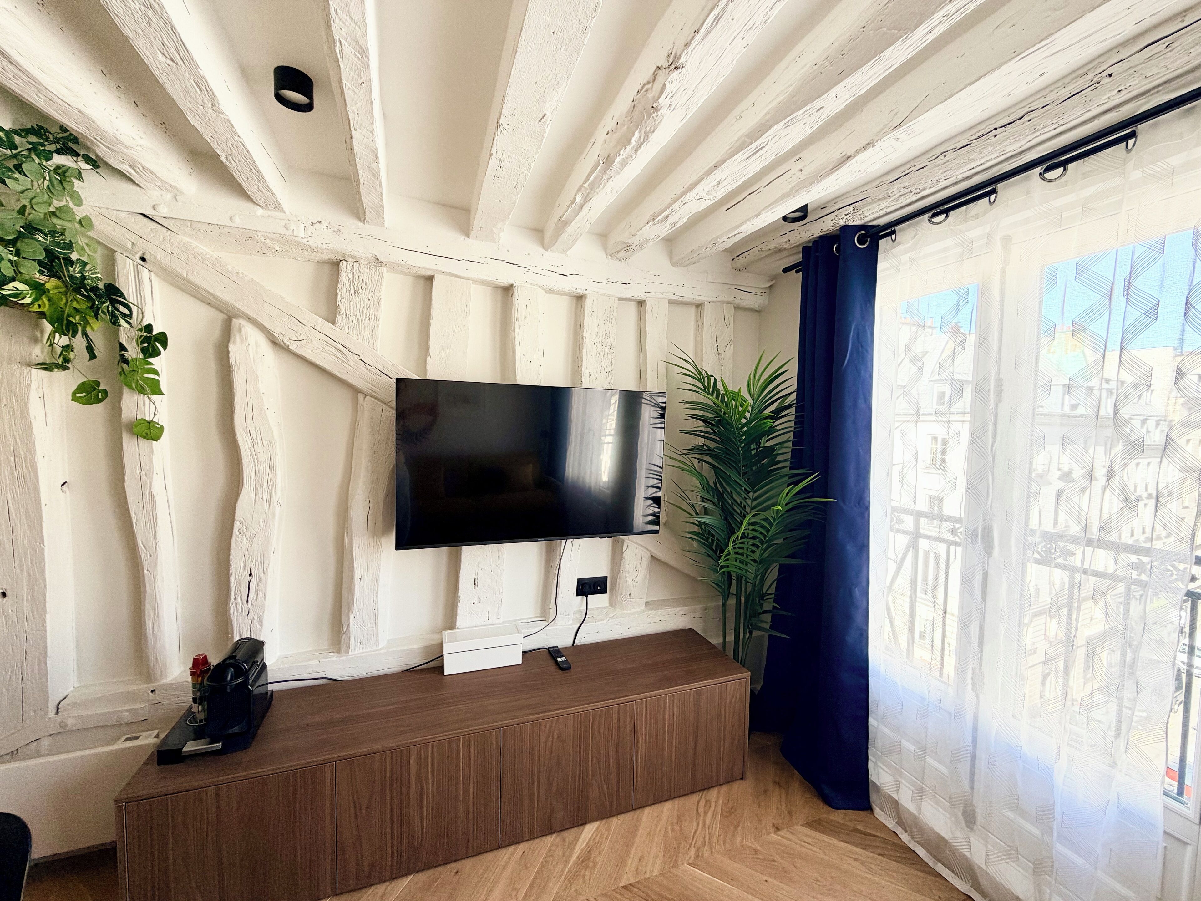 A room with exposed wooden beams, a flat-screen TV mounted on the wall, a wooden bench, and a potted plant.