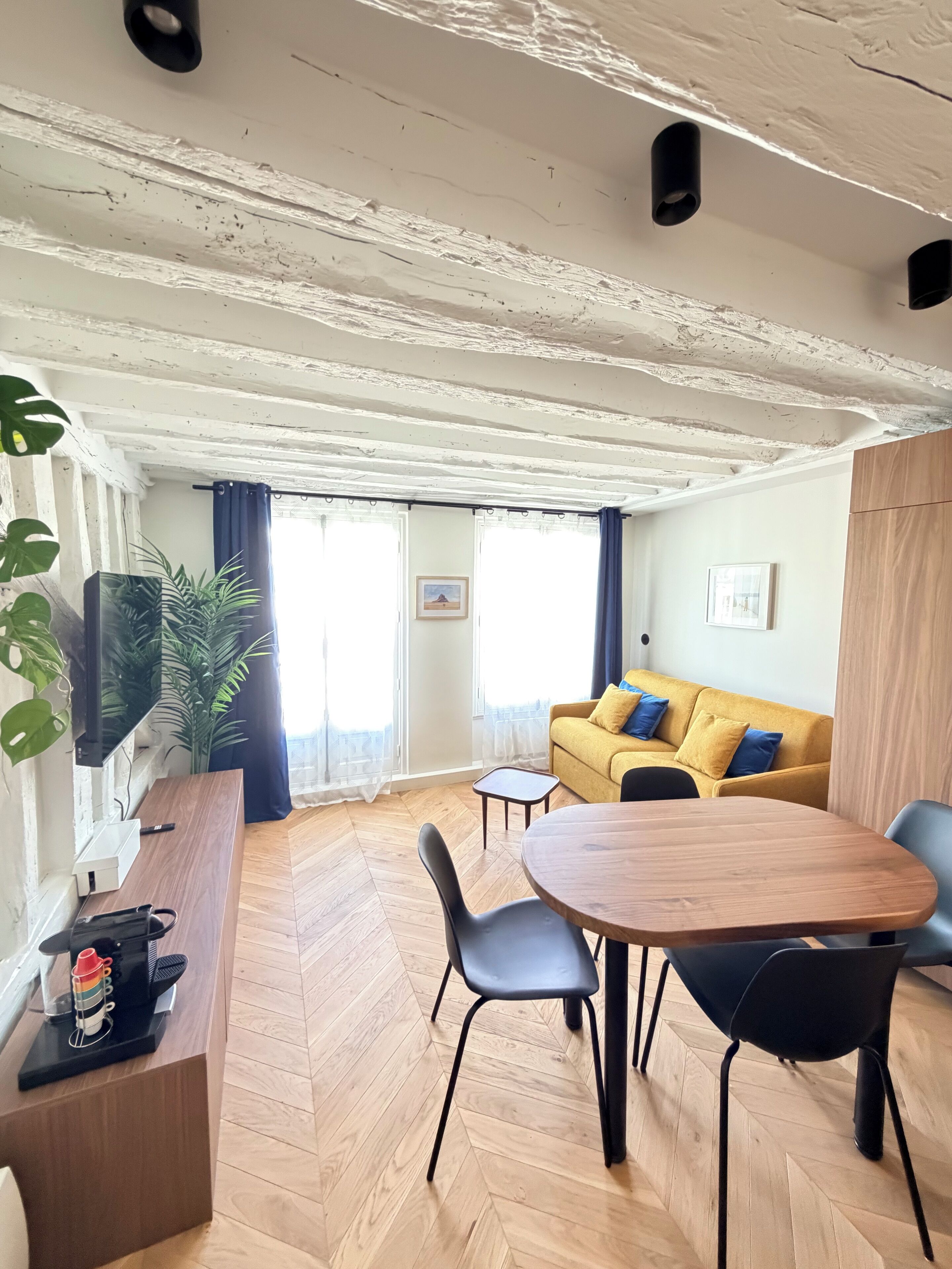 Ein modernes Wohnzimmer mit Holzdielen im Fischgrätmuster, einem runden Holztisch mit Stühlen, einem gelben Sofa mit blauen Kissen und einem großen Fenster mit Vorhängen.