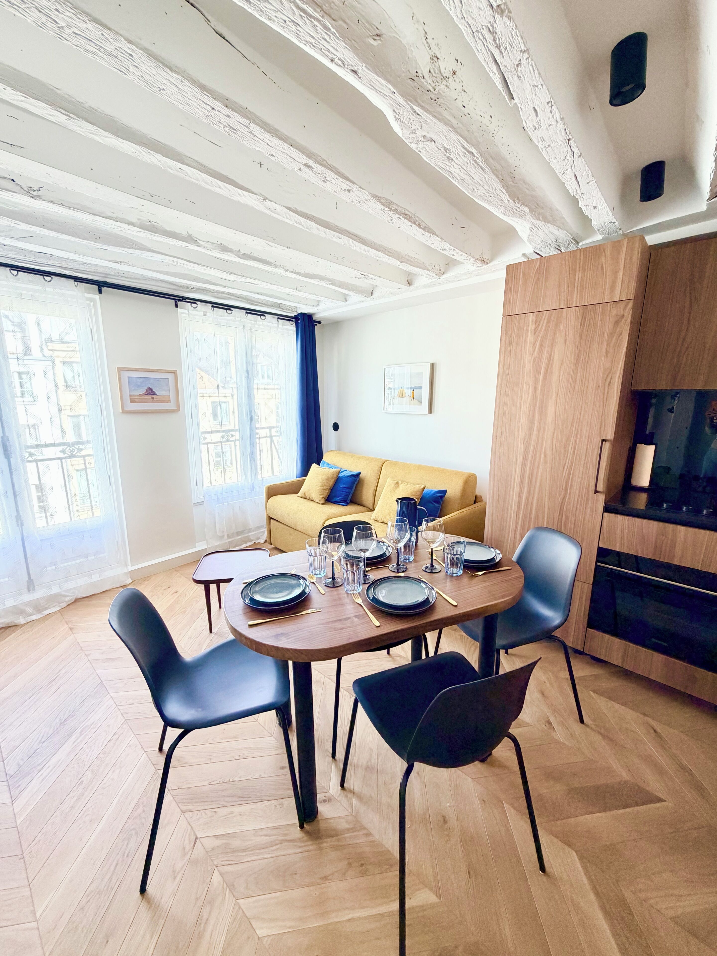 Un comedor con una mesa de madera dispuesta para cuatro personas, rodeada de sillas negras. Un sofá amarillo con cojines azules está apoyado contra la pared, y se aprecia una zona de cocina a la derecha.