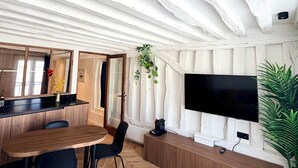 Ein moderner Wohnraum mit Holzdielen im Fischgrätmuster, einem großen Esstisch aus Holz, schwarzen Stühlen, einem an der Wand montierten Flachbildfernseher und einem Blumentopf.
