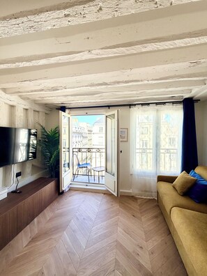 A living room with a herringbone floor, a sofa, a TV, and a balcony with a view of buildings. - Luxury Apartment - Heart of Le Marais with balcony - Amazing location (Paris)