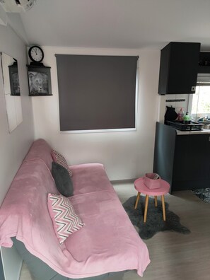 A modern living room with a pink sofa, a grey rug, a small round table, and a kitchen area with cabinets and a window. - Superb location in the Var (Fréjus)