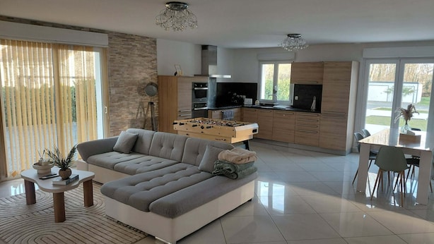 Una sala moderna con un sofá modular, un comedor con mesa y sillas, una cocina con mesa de fútbol, y ventanas grandes que ofrecen luz natural.