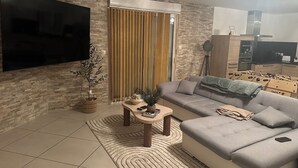 Una sala de estar moderna con un sofá gris, un televisor de pantalla plana montado en una pared de ladrillo, una pequeña mesa de centro de madera y un gran ventanal con persianas verticales.