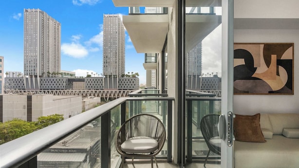 A balcony with a view of tall buildings, a chair, a sofa, and a glass railing. - 501 First | Downtown 1BR | Pool & Skyline Views (Miami)