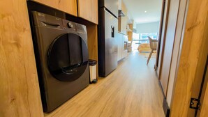 Zona de lavandería con lavadora y secadora, suelo de madera y vista hacia un dormitorio con cama y ventana.