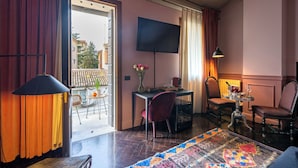 Habitación de hotel con balcón, televisión, mesa de comedor con sillas y alfombra estampada.