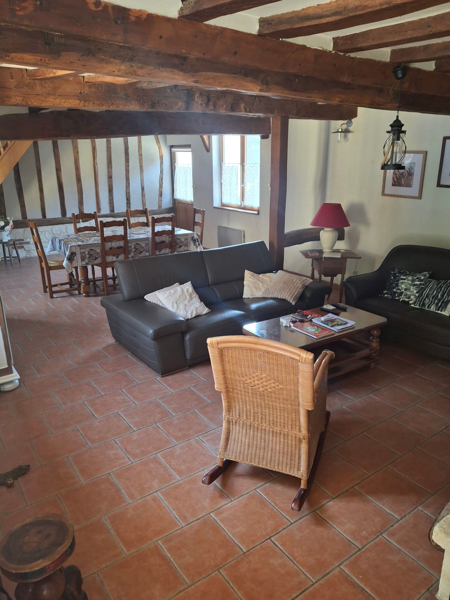 A rustic living room with exposed wooden beams, a black leather sofa, a wicker chair, and a dining area with a table and chairs.