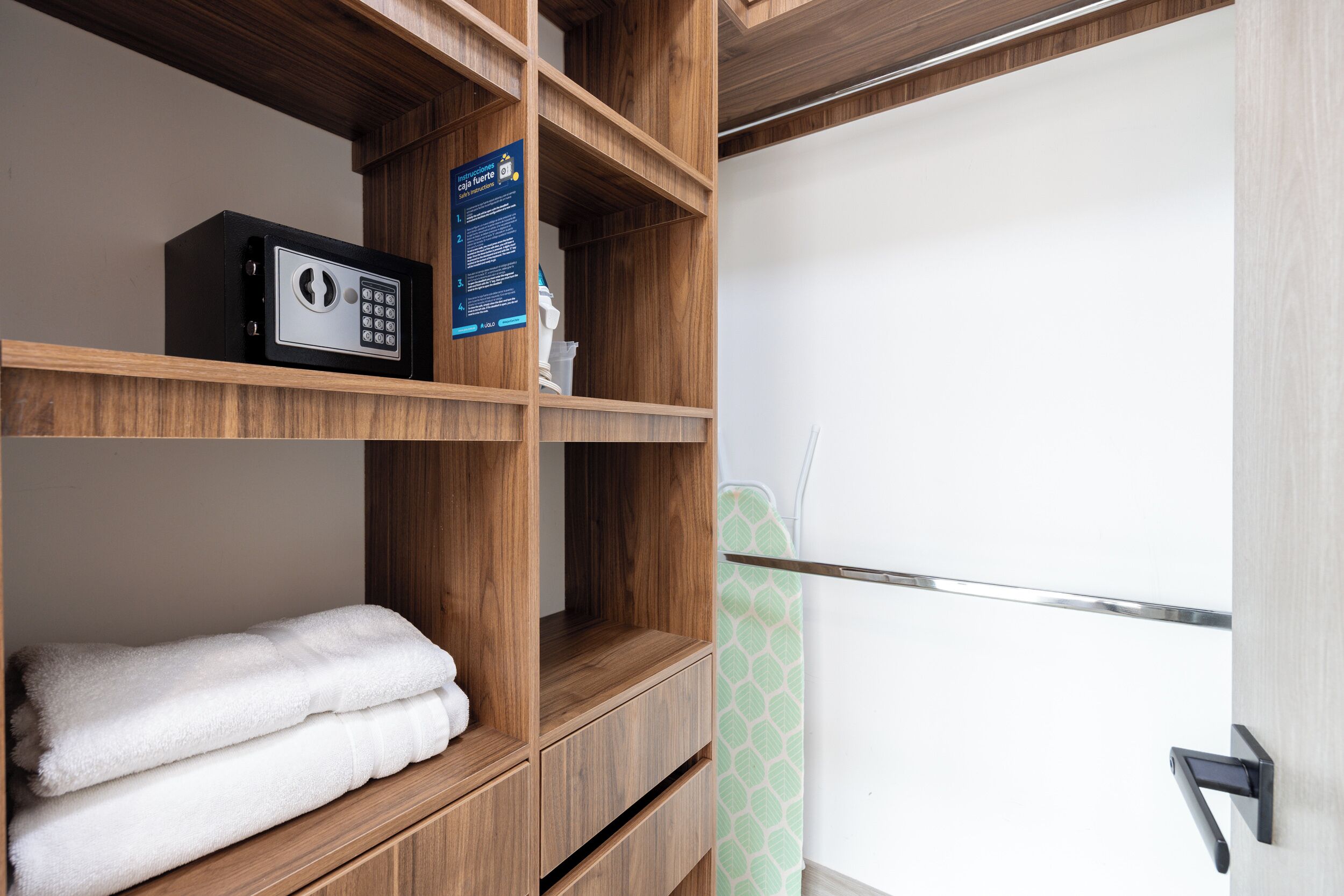 Habitación de hotel con una estantería de madera que contiene una caja fuerte y toallas, una puerta con pomo y una pared con estampado verde.