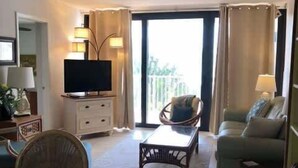 Ein Wohnzimmer mit Fernseher, einem Esstisch, Stühlen, einer Couch und einem Balkon mit Vorhängen.