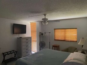 Ein Schlafzimmer mit Bett, Deckenventilator, Fernseher, Kommode und einem Fenster mit Jalousien.