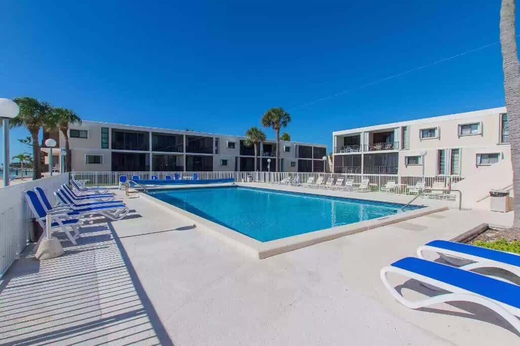 Un área de piscina con sillones reclinables, una piscina azul cristalina y un edificio con balcones.