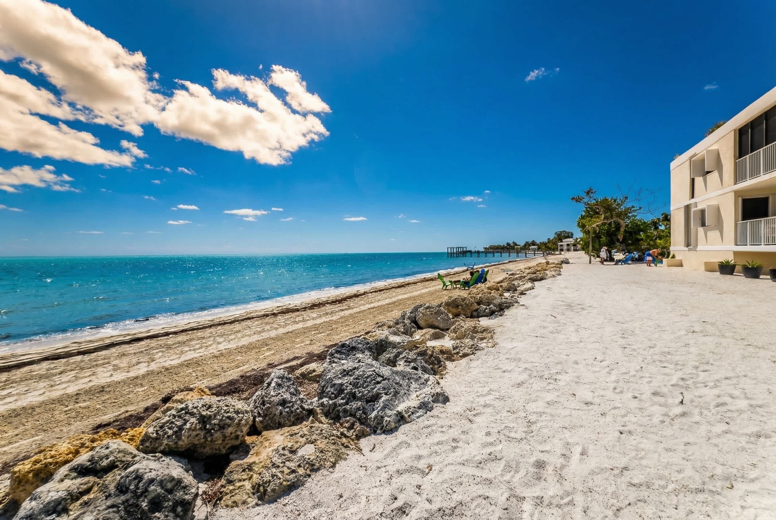 Une promenade en bord de mer, séparée de l’eau par une barrière rocheuse, sous un ciel bleu azur, avec une jetée visible au loin.