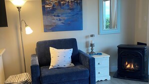 Een woonkamer met een blauwe fauteuil, een schilderij, een vloerlamp, een witte nachtkastje, een spiegel en een open haard.