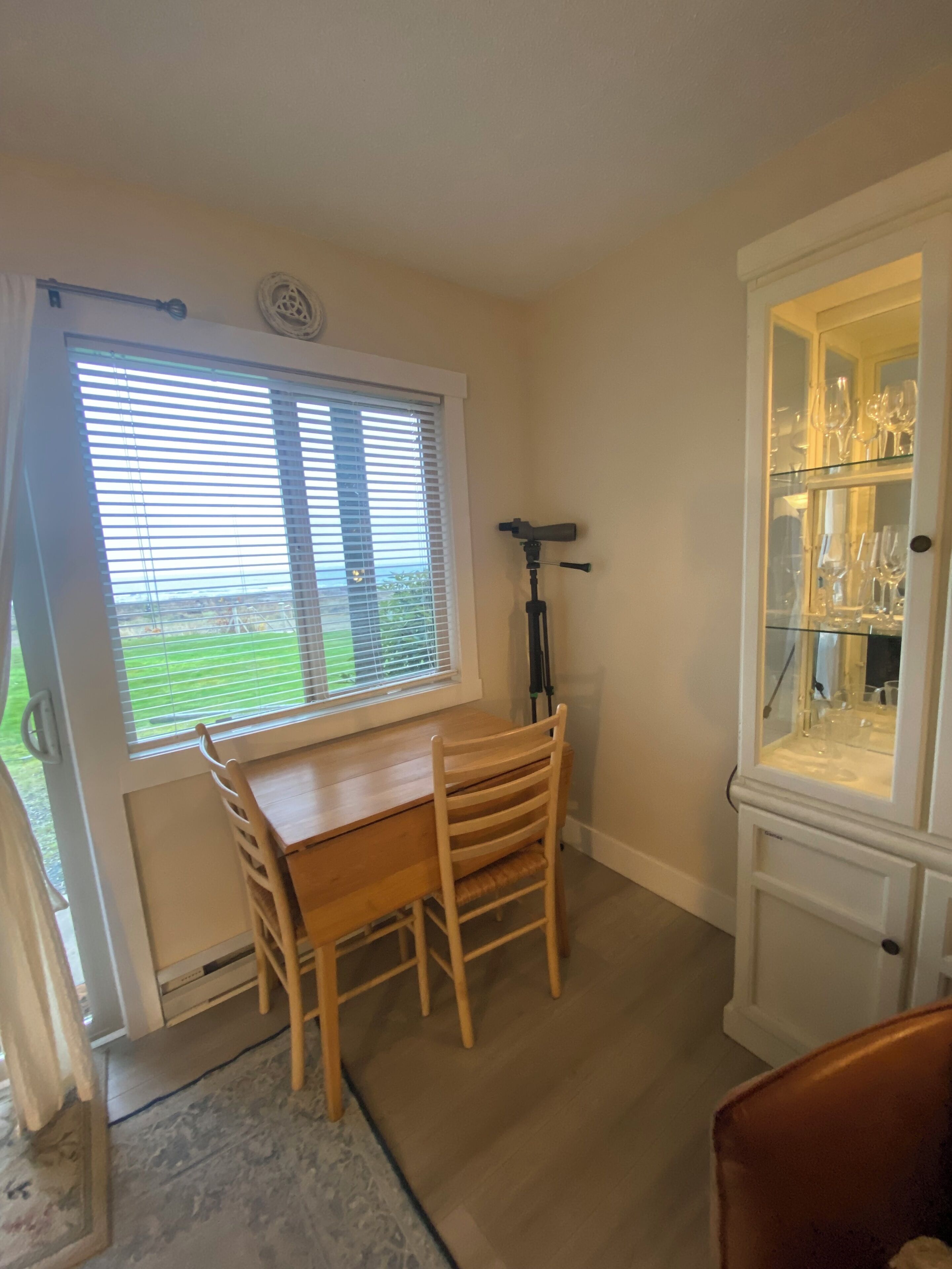 Ein Zimmer mit Holztisch und Stühlen, einem Fenster mit Jalousien, einem Stativ und einer Glasvitrine mit Weingläsern.