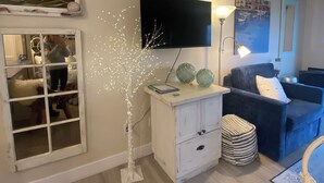 Een woonkamer met een flatscreen televisie, een wit kastje, een blauwe bank en een decoratieve boom met lichtjes.