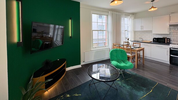 Une cuisine moderne avec des armoires blanches, un four à micro-ondes et une cuisinière. On y trouve une table à manger avec des chaises et un fauteuil vert. Un téléviseur à écran plat est fixé au mur, et deux lampes suspendues sont accrochées au-dessus du plan de travail de la cuisine.