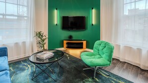 Un salon moderne avec un mur vert, un téléviseur à écran plat, un fauteuil vert, une table basse en verre et une plante en pot.