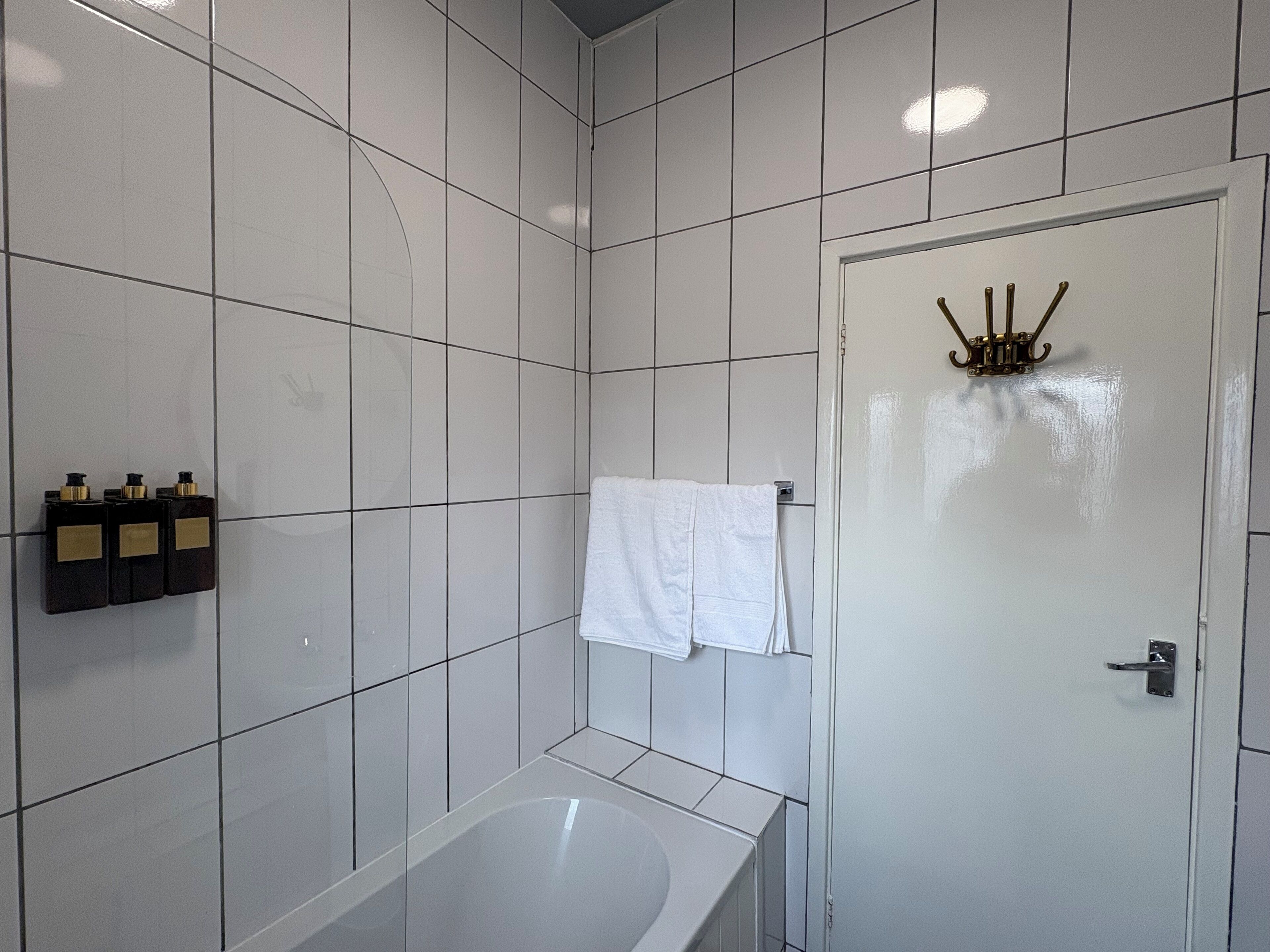 Een badkamer met witte betegelde wanden, een witte badkuip en een deur met een handdoekhouder en flesjes.