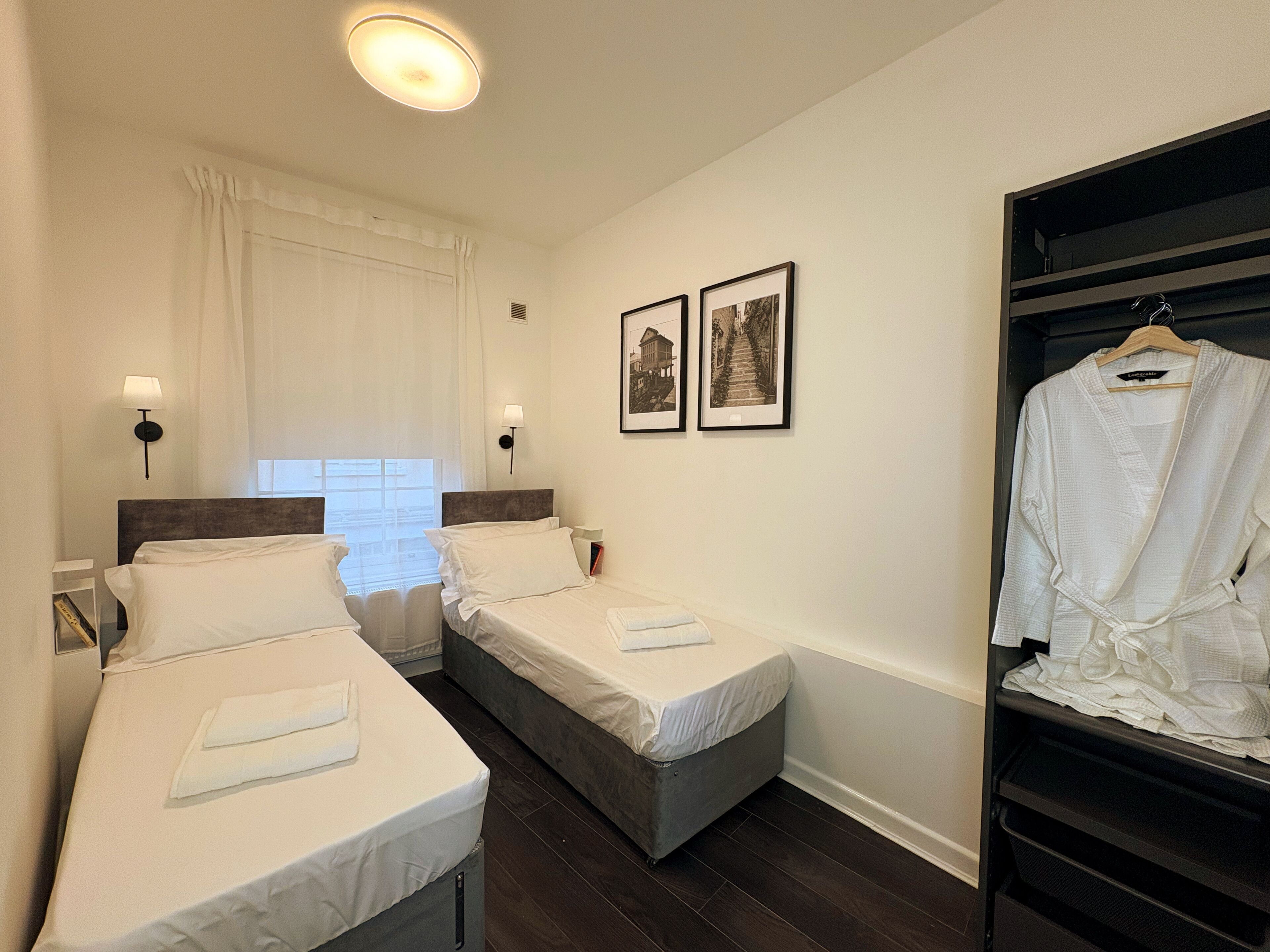 Ein Hotelzimmer mit zwei Einzelbetten, einem Schrank, einem Fenster mit Vorhängen und zwei gerahmten Bildern an der Wand.