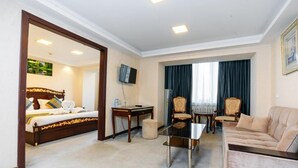 Una habitación de hotel con cama, sofá, una mesita, televisor y ventana con cortinas.